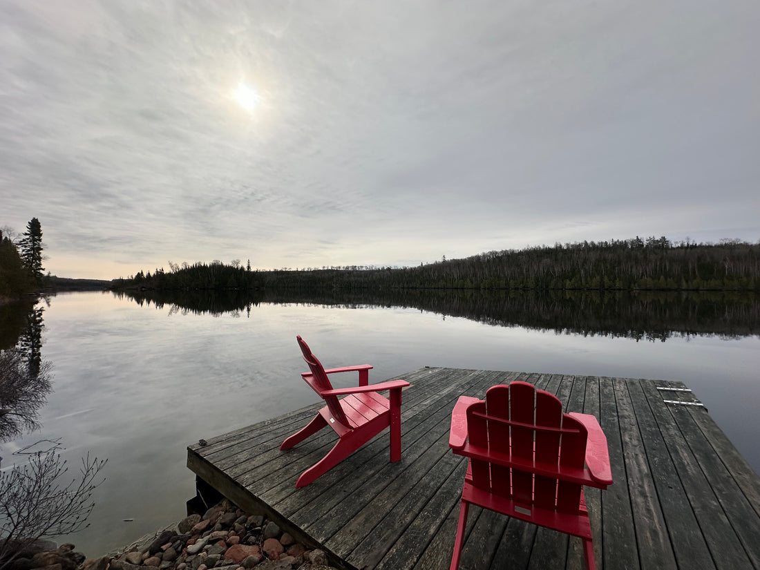 Photo shows two red Adirondack chairs on a wooden dock. The day is cloudy and the sun is barely showing through the clouds. You can see a silhouette of the tree line. The lake is calm like glass and reflects the grey hues of the sky.